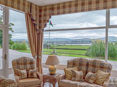 A sitting area with armchairs cushions a lamp and large windows overlooking a green landscape and hills at Tal Gwynedd in Dwyran near Newborough