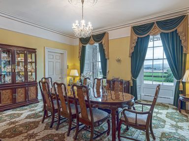A dining room with a wooden table and chairs a china cabinet and large windows with curtains at Tal Gwynedd in Dwyran near Newborough