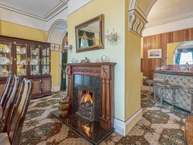 A dining room with wooden chairs a glass cabinet a wood fireplace and a bar with stools at Tal Gwynedd in Dwyran near Newborough