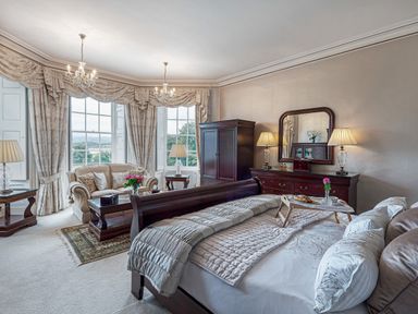 A bedroom with a bed a sofa a wooden wardrobe a dresser with lamps and a large window with curtains at Tal Gwynedd in Dwyran near Newborough