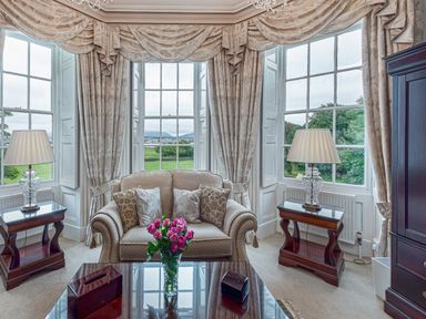A sitting area with a loveseat sofa and decorative pillows between three large windows with draped curtains and two side tables with lamps at Tal Gwynedd in Dwyran near Newborough