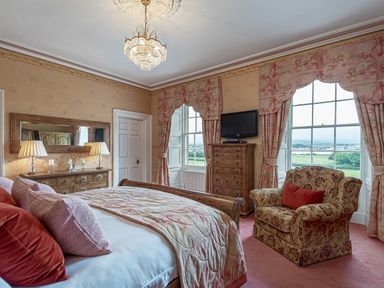 A bedroom with a bed a wooden dresser with lamps a mirror an armchair a chest of drawers with a television and two large windows with curtains at Tal Gwynedd in Dwyran near Newborough