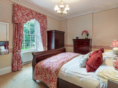 A bedroom with a wooden bed chest of drawers wardrobe red patterned curtains and matching bedding at Tal Gwynedd in Dwyran near Newborough