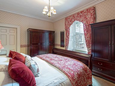 A bedroom with a bed with red pillows and blanket two dark wooden wardrobes and a window with red curtains at Tal Gwynedd in Dwyran near Newborough