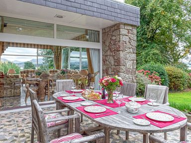 An outdoor dining table set with plates glasses and flowers next to a stone wall and glass doors with garden and trees in the background at Tal Gwynedd in Dwyran near Newborough