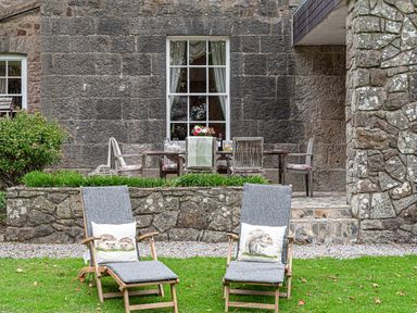 Two cushioned wooden lounge chairs with animal-themed pillows on grass in front of a stone patio with a dining table and chairs at Tal Gwynedd in Dwyran near Newborough