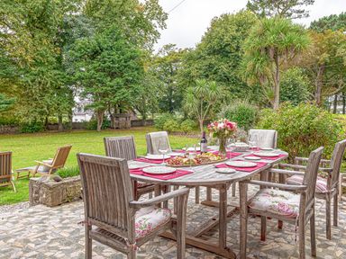 A wooden outdoor dining table set with plates glasses flowers and a bottle of wine surrounded by wooden chairs on a stone patio with trees and grass at Tal Gwynedd in Dwyran near Newborough