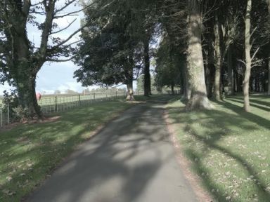 A paved path surrounded by grass and trees with a field and fence visible in the background at Tal Gwynedd in Dwyran near Newborough