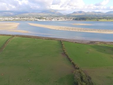 A landscape with green fields and sheep near a river with sandbanks and a town in the background with hills beyond at Tal Gwynedd in Dwyran near Newborough