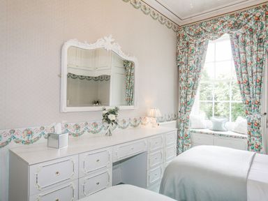 A bedroom with twin beds a white dresser a large mirror floral curtains and a window seat at Tal Gwynedd in Dwyran near Newborough