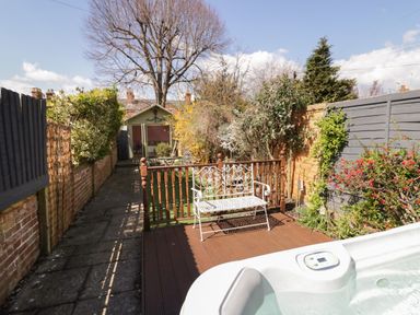 A backyard with a hot tub a white metal bench wooden deck paved pathway and a garden shed at Heath Cottage in Stratford-Upon-Avon