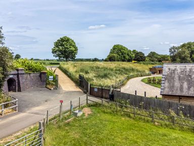 An outdoor rural scene with a gated entrance, gravel driveway, brick structure, grassy fields, and a wooden building at Willow Lodge At Bridge Lake Farm & Fishery in Chacombe