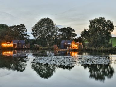 A lake with two small cabins and trees reflected in the water at Willow Lodge At Bridge Lake Farm & Fishery in Chacombe