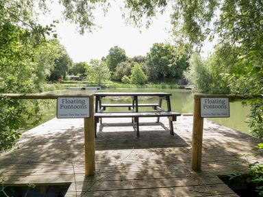 A wooden deck with a bench and signs by the water at Willow Lodge At Bridge Lake Farm &amp; Fishery Chacombe