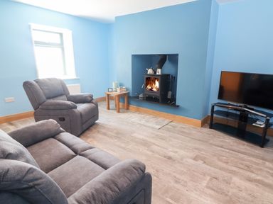 A living room with gray sofas a wood burning stove a small wooden table and a television on a stand at Lack Cottage Annascaul near Camp County Kerry