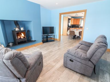 A living room with two gray recliner chairs a wood stove and a TV facing a dining area with a table and chairs at Lack Cottage Annascaul near Camp County Kerry