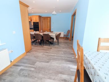 A dining area with a long table set with plates and mugs surrounded by chairs and a kitchen with wooden cabinets at Lack Cottage in Annascaul near Camp County Kerry