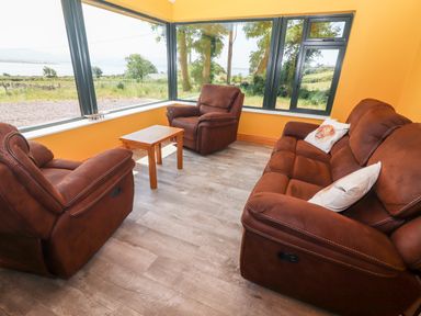 A sitting room with brown armchairs sofa and a wooden table by large windows at Lack Cottage Annascaul near Camp County Kerry