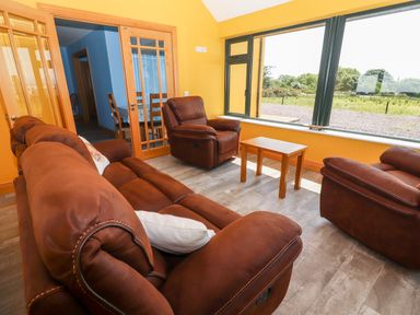 A living room with brown sofas and armchairs around a wooden table beside large windows at Lack Cottage Annascaul near Camp County Kerry