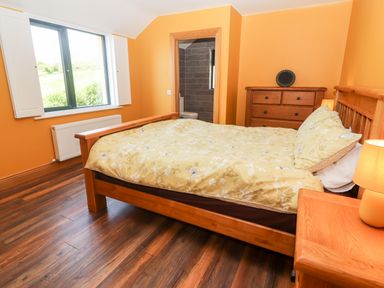 A bedroom with a wooden bed and floral bedding a wooden dresser and nightstand with a lamp and an open door to a bathroom at Lack Cottage Annascaul near Camp County Kerry