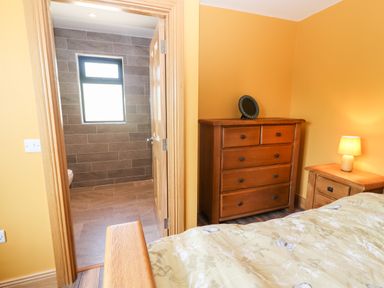 A bedroom with wooden furniture including a chest of drawers and bedside table with a lamp and an open door leading to a tiled bathroom at Lack Cottage Annascaul near Camp County Kerry