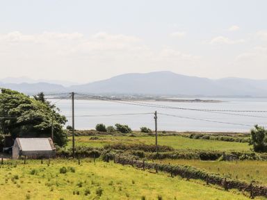 A rural landscape with green fields, a small stone building, power lines, and mountains by water at Lack Cottage in Annascaul near Camp, County Kerry
