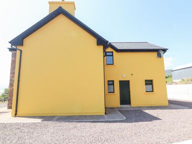 The exterior side and back of a yellow house with a black door and windows and a gravel driveway at Lack Cottage Annascaul near Camp County Kerry