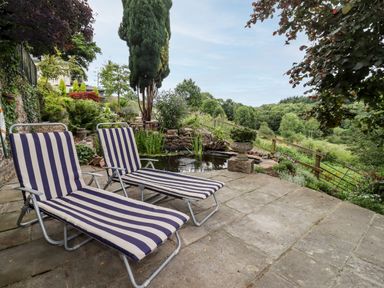 An outdoor patio with two striped lounge chairs by a small pond and a wooden fence with trees in the background at The Tack Room in Lydney