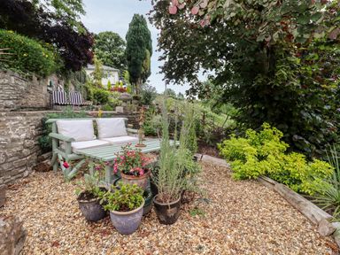 An outdoor seating area with a green wooden bench and table surrounded by potted plants and gravel ground at The Tack Room in Lydney