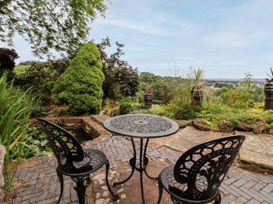 A metal outdoor table with two chairs on a brick patio overlooking a garden with shrubs and a distant view at The Tack Room in Lydney