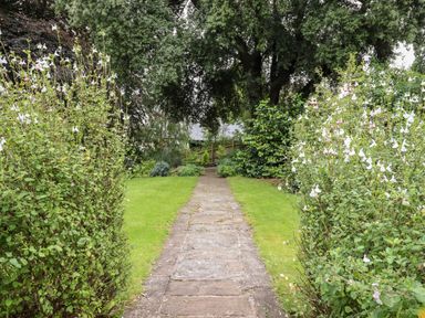 A stone pathway through a garden with grass and flowering shrubs at The Tack Room in Lydney