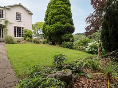 A garden with trimmed lawn shrubs large trees and a white house at The Tack Room in Lydney