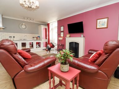 A living room with two red leather chairs a small red table with a plant a wall-mounted TV and a kitchenette at The Tack Room in Lydney
