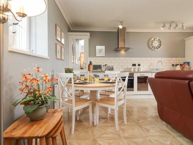 A kitchen and dining area with a round wooden table set with plates and glasses near a red leather sofa at The Tack Room in Lydney