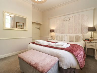 A bedroom with a bed with white and pink bedding bedside tables with lamps a mirror on the wall and a pink cushioned bench at The Tack Room in Lydney