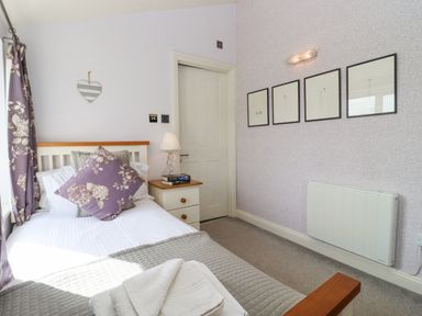 A bedroom with a single bed with floral pillows beside a bedside table with a lamp and books and framed pictures on the wall at The Tack Room in Lydney