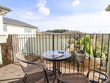 A balcony with a round glass table, two wicker chairs, a magazine, sunglasses, two glasses, and a champagne bottle in a bucket overlooking trees and hills at The Tack Room in Lydney