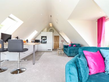An open plan room with a kitchen area including stools and appliances and a sitting area with teal sofas and pink cushions at Higher Kernick View in Helland near Blisland