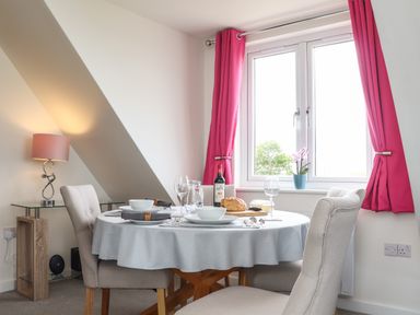 A dining area with a round table set for four near a window with pink curtains at Higher Kernick View in Helland near Blisland