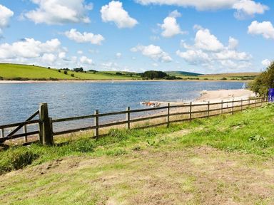 A grassy area with a wooden fence overlooking a body of water with boats and hills in the background at Higher Kernick View in Helland near Blisland
