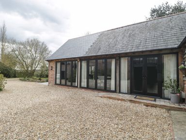 An exterior view of a building with large black-framed windows and a gravel driveway at The Courtyard - Hilltop Barn in Winterborne Zelston near Wareham