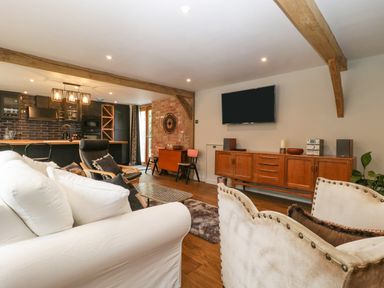 An open plan living room and kitchen with wooden beams a white sofa a black armchair and a wooden sideboard with a wall mounted TV at The Courtyard - Hilltop Barn in Winterborne Zelston near Wareham