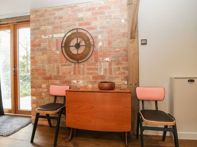 A brick wall with a wooden clock above a wooden table and two chairs with pink backs in a room at The Courtyard - Hilltop Barn in Winterborne Zelston near Wareham