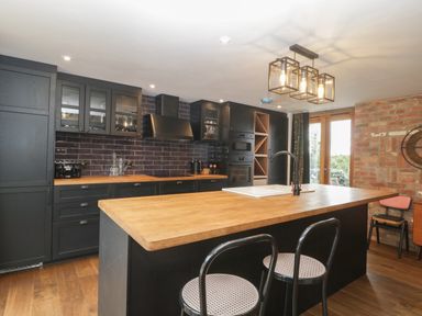 A kitchen with black cabinets wooden countertops an island with a sink two chairs a brick wall and glass doors at The Courtyard - Hilltop Barn in Winterborne Zelston near Wareham