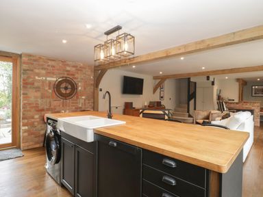 An open kitchen and living area with a wooden island countertop black cabinets a white farmhouse sink and a washing machine at The Courtyard - Hilltop Barn in Winterborne Zelston near Wareham