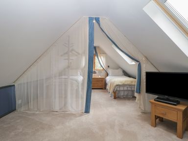 An attic bedroom with two single beds separated by sheer curtains a wooden nightstand and a television on a wooden stand at The Courtyard - Hilltop Barn in Winterborne Zelston near Wareham