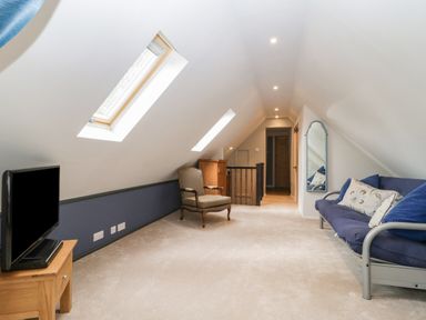 A narrow attic room with two skylights a TV on a wooden stand an armchair a chest of drawers a blue sofa with cushions and a wall mirror at The Courtyard - Hilltop Barn in Winterborne Zelston near Wareham