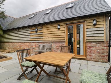 An outdoor patio with wooden table and chairs with cushions and a bench against a wooden and brick building at The Courtyard - Hilltop Barn Winterborne Zelston near Wareham