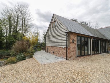 A building with brick and wood siding with glass doors on a gravel driveway with trees and plants nearby at The Courtyard - Hilltop Barn in Winterborne Zelston near Wareham