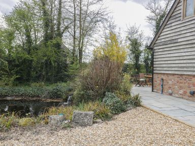 A gravel path and paved patio next to a wooden house with a small pond surrounded by plants and trees at The Courtyard - Hilltop Barn in Winterborne Zelston near Wareham